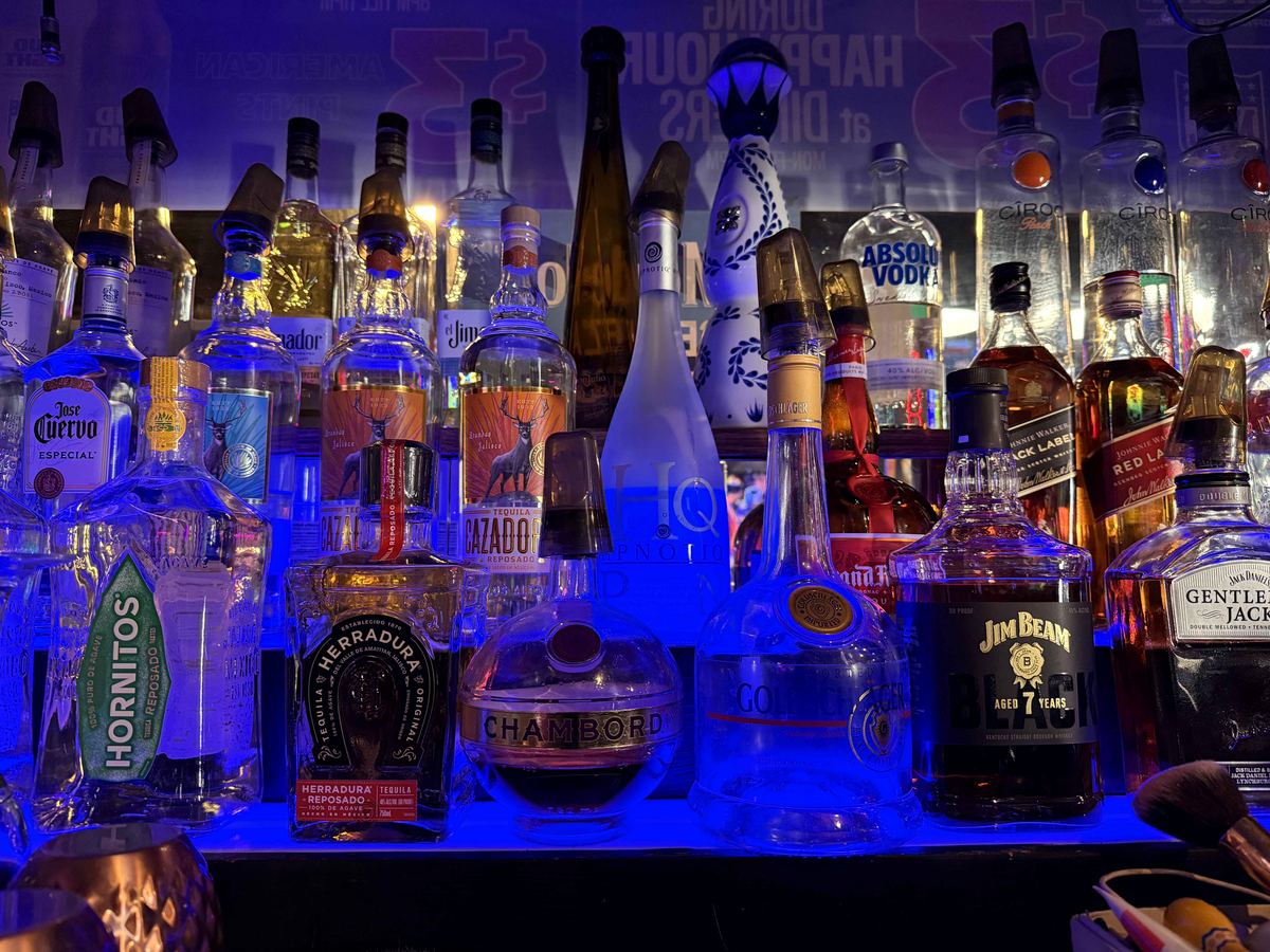 Back bar at Dingers Sports Bar illuminated by electric cobalt blue LED lighting. Multiple spirits bottles including Hornitos, Herradura, Patron, Jim Beam visible on shelves.