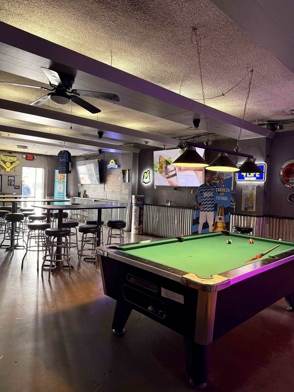 Pool room at Dingers Sports Bar at night. Back wall shows IPA red neon sign, TUF yellow neon sign, projector mount visible on the ceiling. One pool table in foreground.