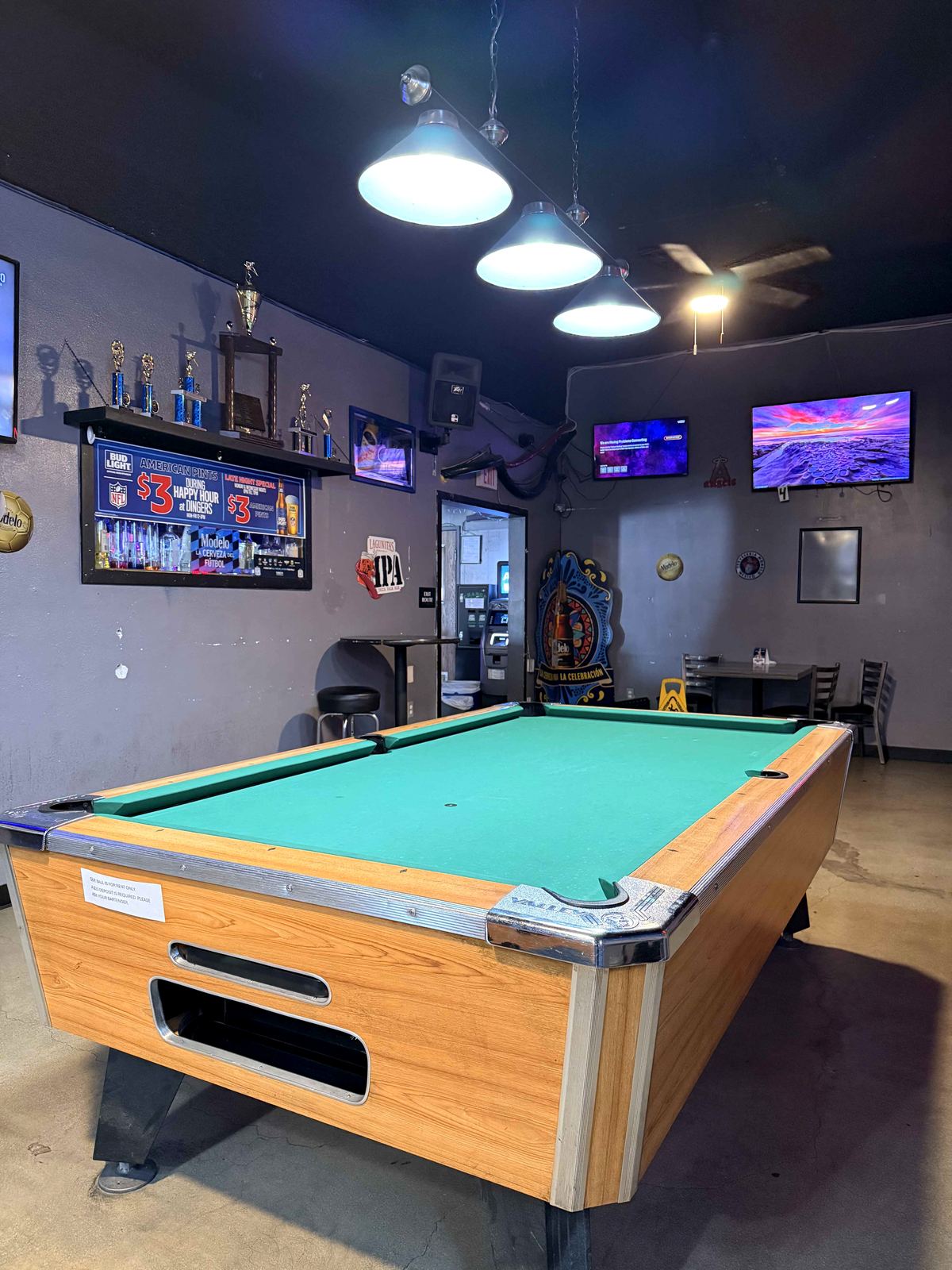 Pool league trophy shelf at Dingers Sports Bar. Multiple gold and silver trophies on the shelf, IPA red neon sign visible beside them, happy hour chalkboard in the background.
