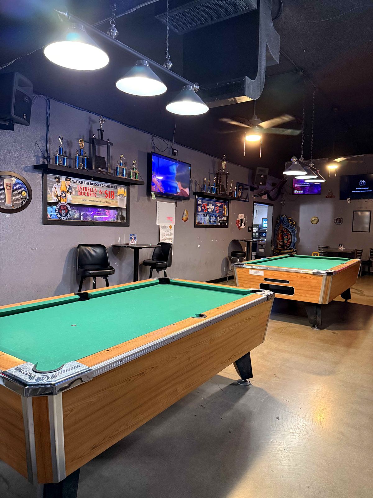 Two pool tables at Dingers Sports Bar. Trophy shelf on the left wall. TVs in background showing a purple-hued sports broadcast.